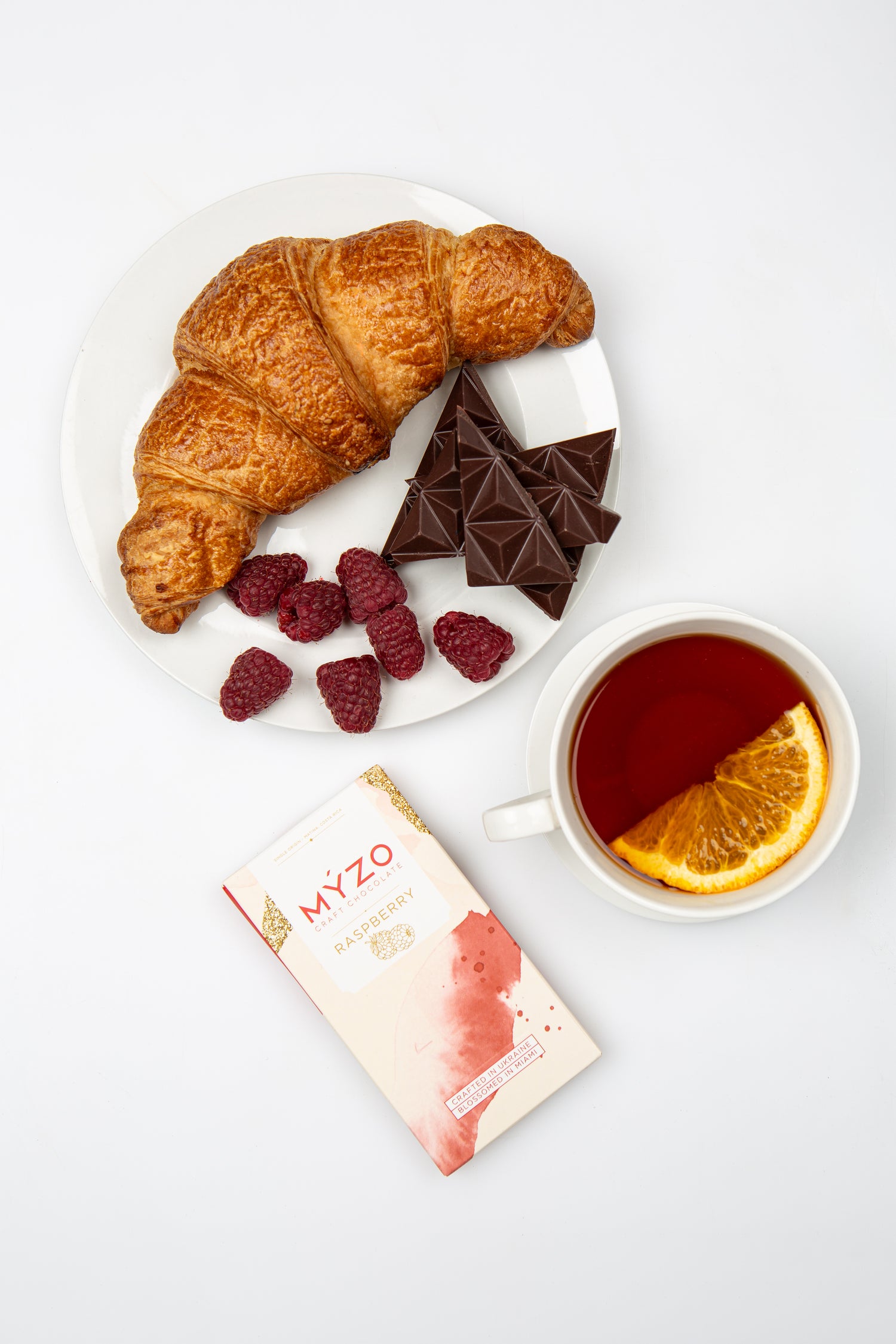 Plate of croissants, chocolate bars, raspberries, and a cup of tea with an orange slice on a white background.